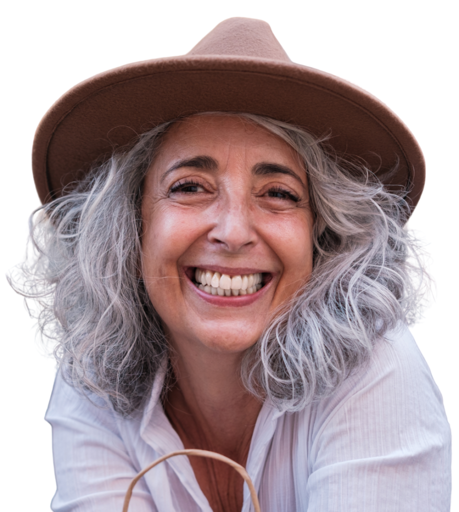 Smiling older woman with shoulder-length gray hair, wearing a wide-brimmed brown hat and a white shirt, looking directly at the camera against a white background, confidently embracing life despite experiencing hearing loss.