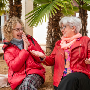 Two older women wearing red jackets and glasses sit under palm trees, smiling and laughing while having a lively conversation outdoors.