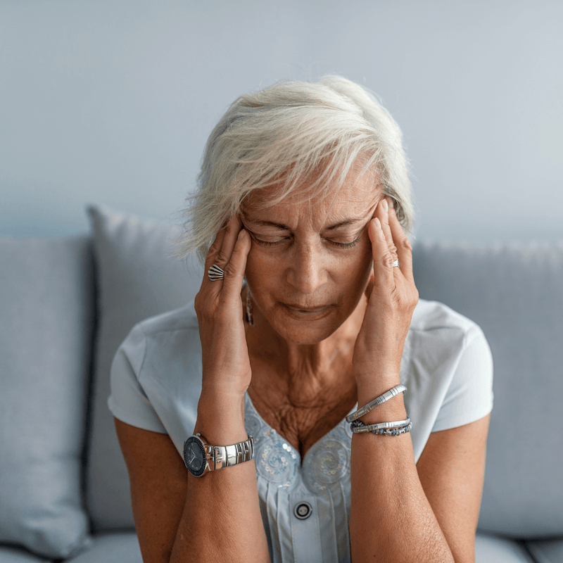 An older woman sitting on a sofa with her eyes closed, holding her hands to her temples as if in pain.