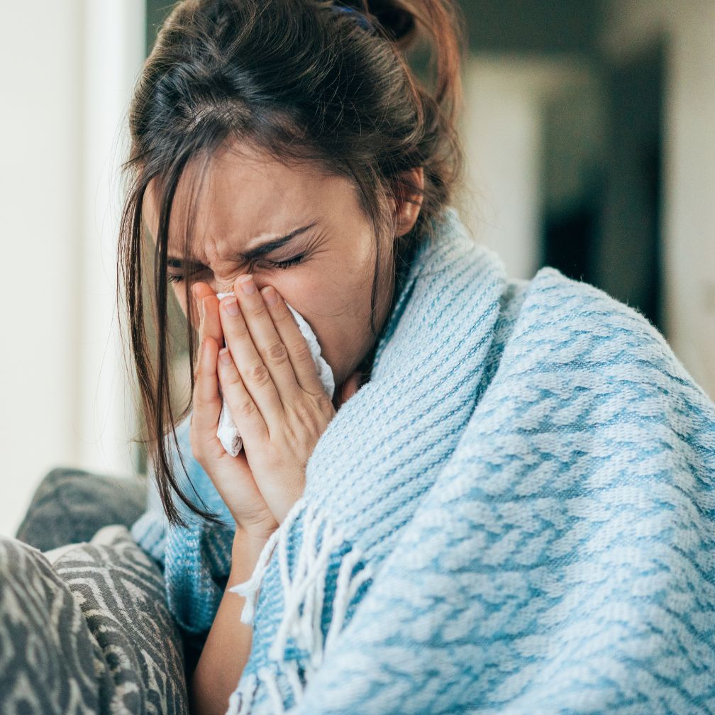 A woman wrapped in a blue blanket sneezes into a tissue.