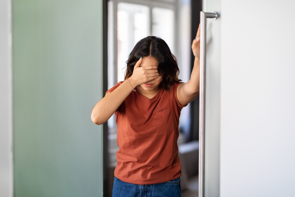A woman in a rust-colored t-shirt leans against a door frame with her hand over her eyes.